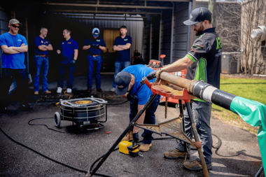 APS instructor demonstrating trenchless pipe repair technique to a group of contractors during hands-on APSTA training session with real pipe and drain cleaning equipment