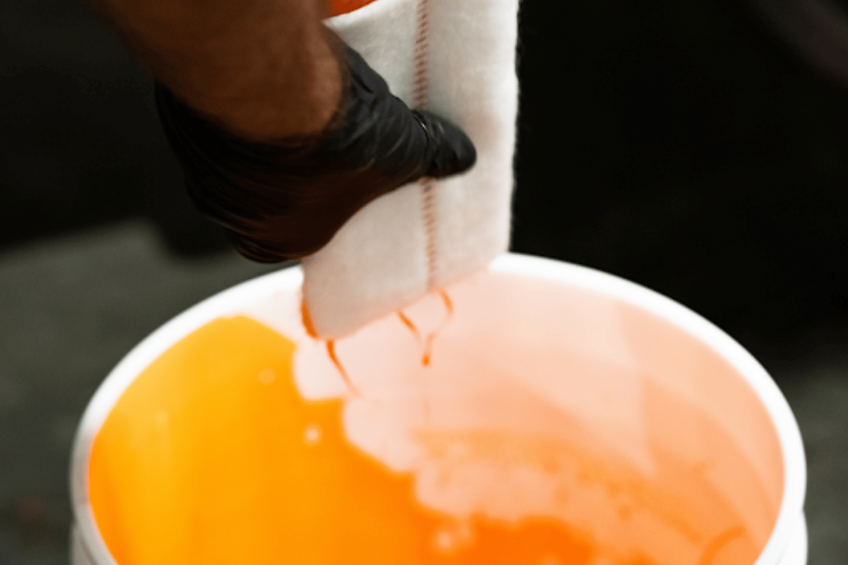 Close-up of a gloved hand squeezing excess orange resin from a trenchless CIPP liner sample into a white bucket during the liner wet-out process.